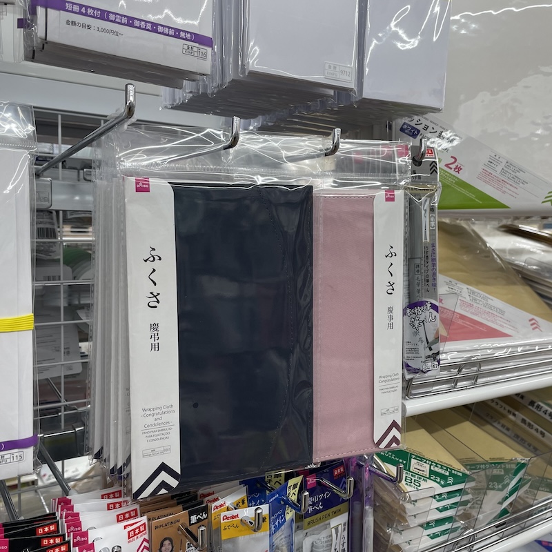 ダイソーの売り場で撮影したふくさの陳列。左に紺色の慶弔用、右にピンクの慶事用が吊り下げられ、周囲に金封や筆ペンが並んでいる